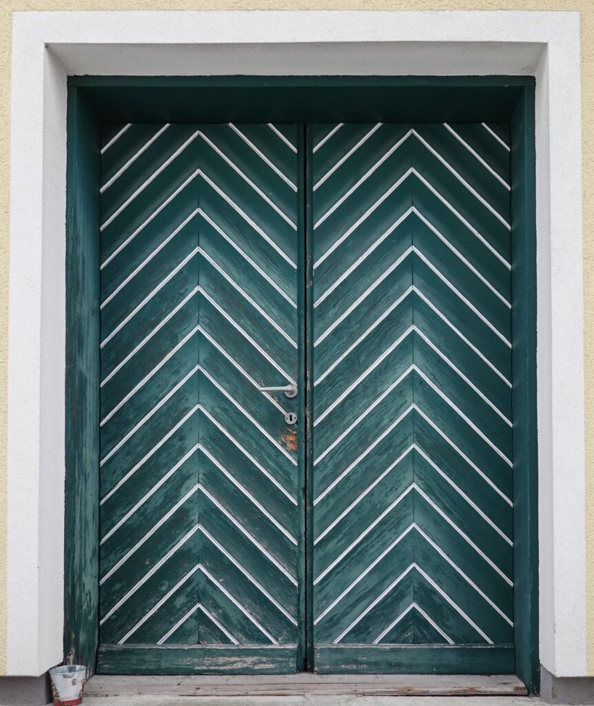 Green door featuring a chevron pattern with rustic texture on a white frame wall.