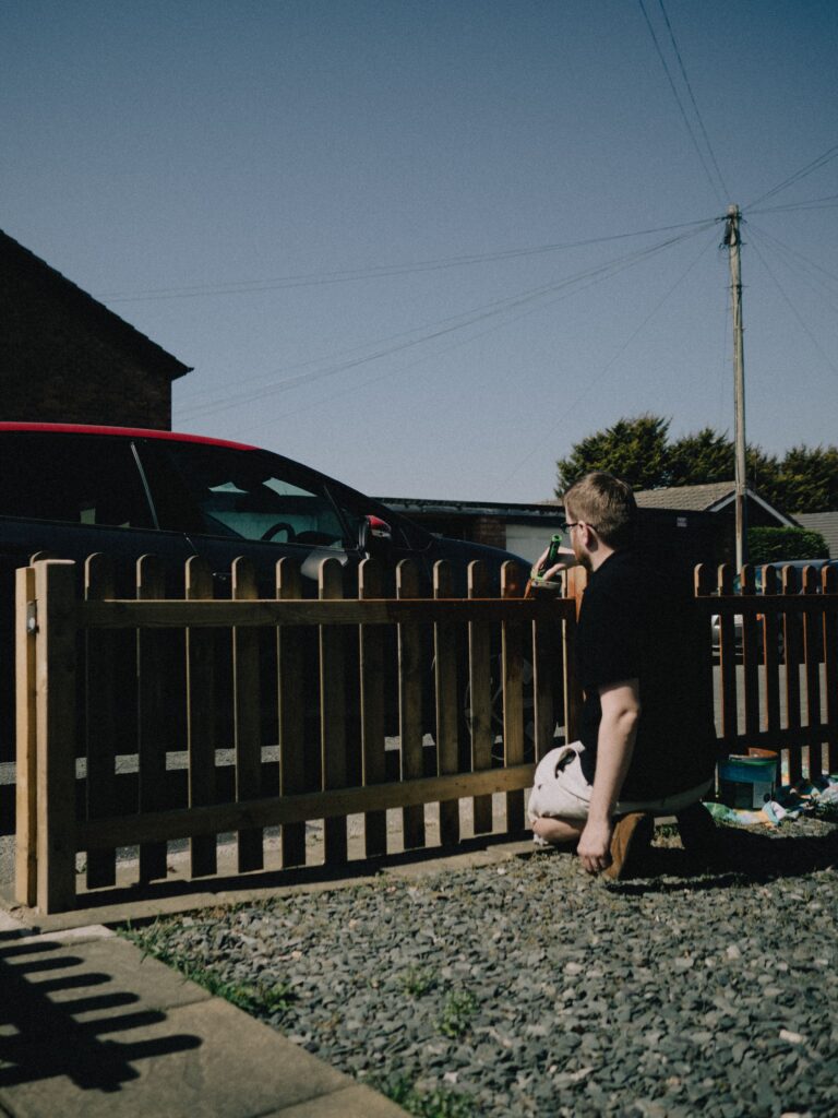A man painting a wooden fence with black paint on a bright summer day.